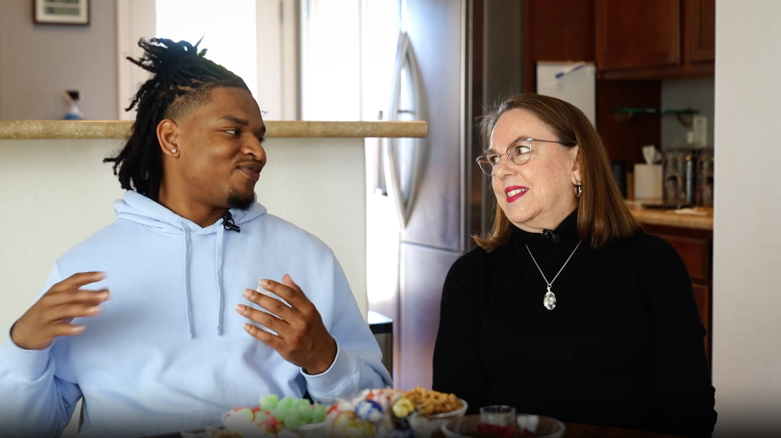 A mother and her grown son in mid-conversation at the kitchen counter, captured during an interview.